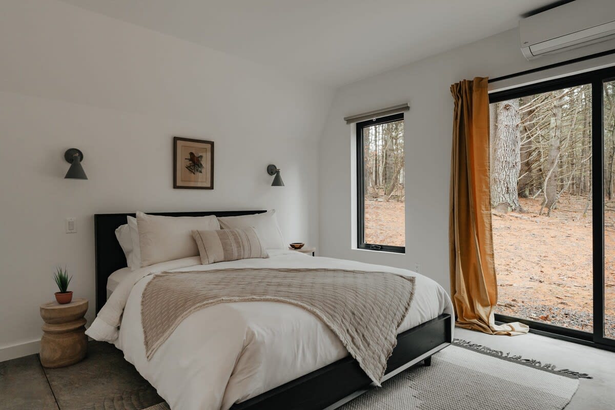 A bright bedroom with a black-framed bed dressed in white and beige linens, flanked by two dark wall sconces. A large glass door with mustard yellow curtains and a tall window offer views of a dense wooded area.