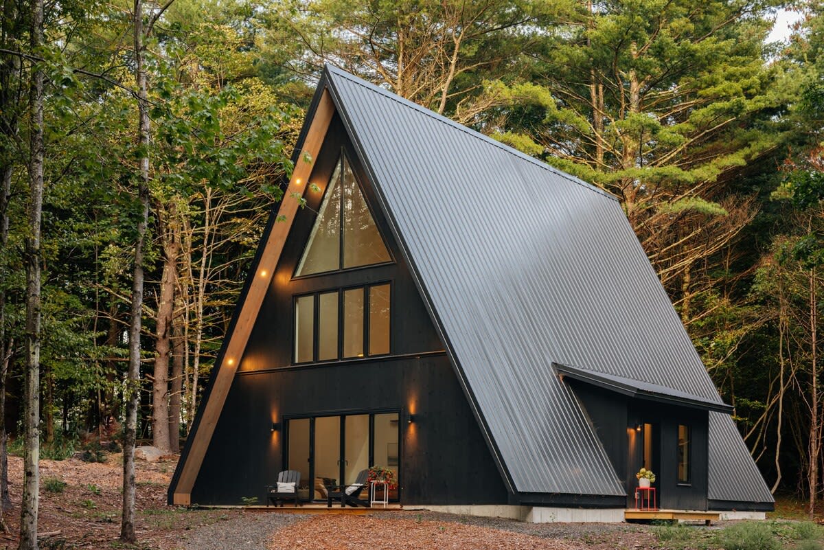 Contemporary black A-frame cabin with a dark metal roof and large glass windows, situated in a lush green forest. A small deck with outdoor seating is visible at the front entrance.
