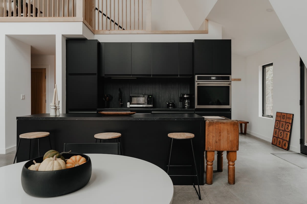 A sleek black kitchen features flat-panel cabinetry, a large island with a dark countertop, and wooden bar stools, positioned next to a round white dining table. A rustic wooden butcher block adds a touch of warmth.