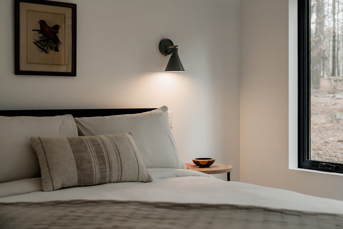 A close-up of a cozy bedroom corner featuring a bed with white and striped pillows, a small round wooden side table with a decorative bowl, and a dark wall sconce providing ambient light. A window offers a glimpse of the outdoors.