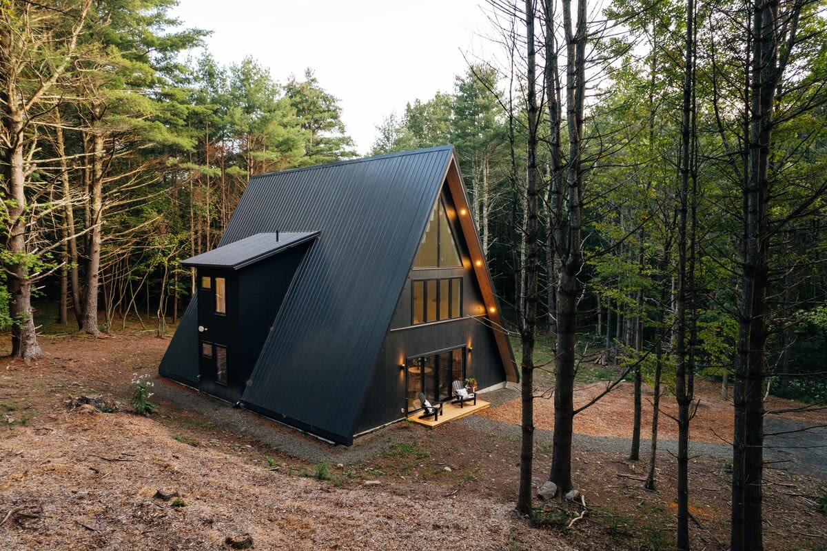 Elevated view of the black A-frame cabin with its dark metal roof and prominent windows, nestled among tall pine trees. The surrounding forest floor is covered in natural debris, suggesting a secluded location.