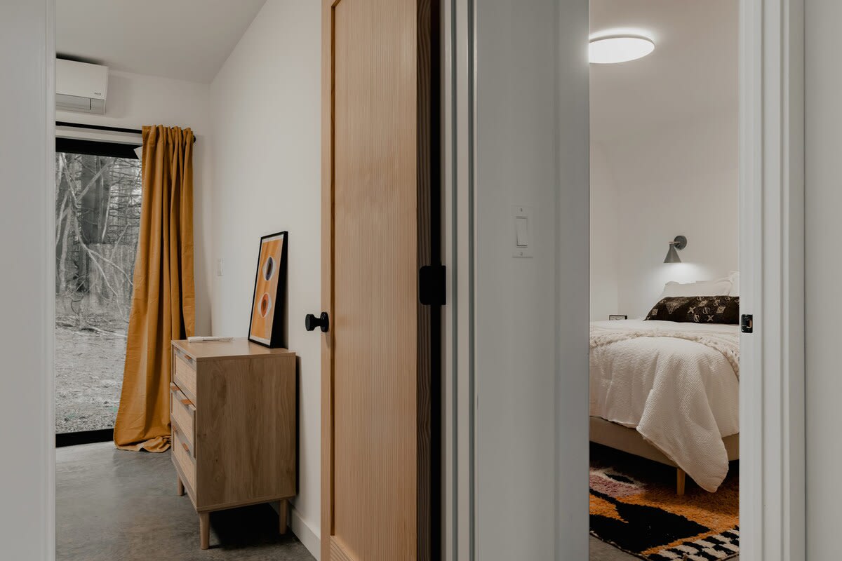 A view from a hallway into a bedroom with a light wood dresser, a framed abstract print, and a large glass door with mustard yellow curtains leading to a wooded outdoor area. Through an open doorway, another bedroom with a bed and a round ceiling light is visible.