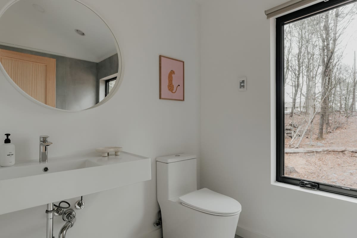 Bright white bathroom with a modern floating sink, a round mirror, and a white toilet. A large window provides a view of a wooded area, and a framed leopard print art piece adds a touch of color.
