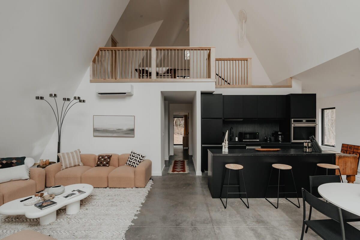A modern open-concept living space features a light peach sectional sofa on a textured rug, facing a white organic-shaped coffee table, with a black kitchen island and dining area visible beyond. A wooden railing loft overlooks the space.