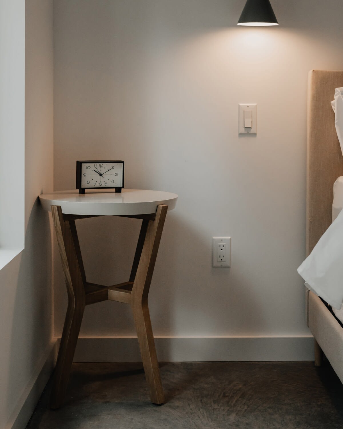 A bedside vignette in a bedroom, featuring a white round side table with wooden legs, a black alarm clock, and a wall outlet, set against a light-colored wall with a beige upholstered headboard.