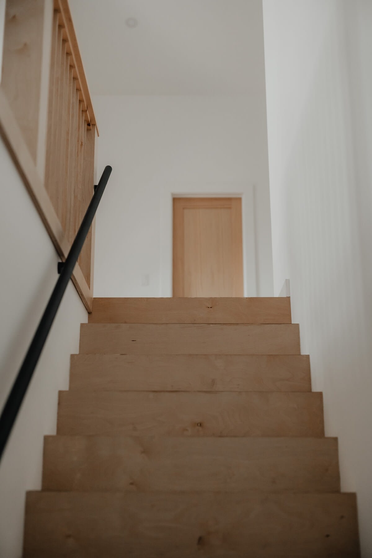 A minimalist wooden staircase with light wood treads and a black metal handrail, leading upwards towards a landing with a light wood door visible in the distance.