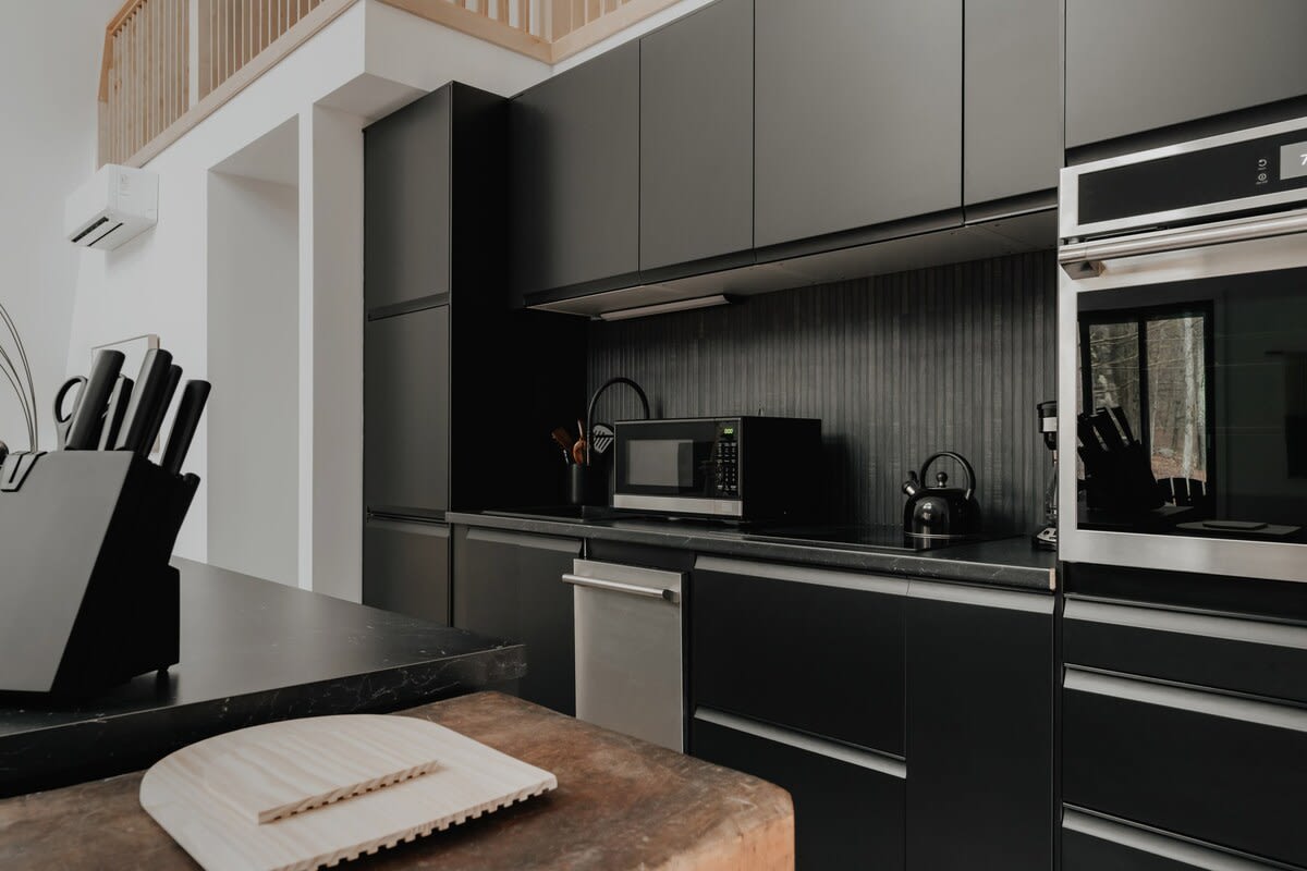 A modern black kitchen counter showcases a built-in oven, microwave, and dishwasher, with sleek black cabinetry above and below. A wooden cutting board rests on a rustic wooden block in the foreground.