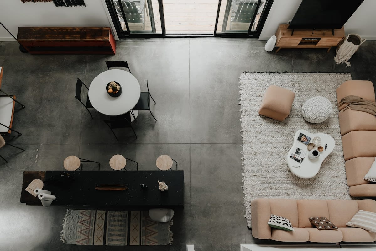 An aerial view reveals the open-concept layout with a light peach sectional sofa on a textured rug, a white organic coffee table, and a black kitchen island with bar stools. A round dining table and large glass doors leading outside are also visible.