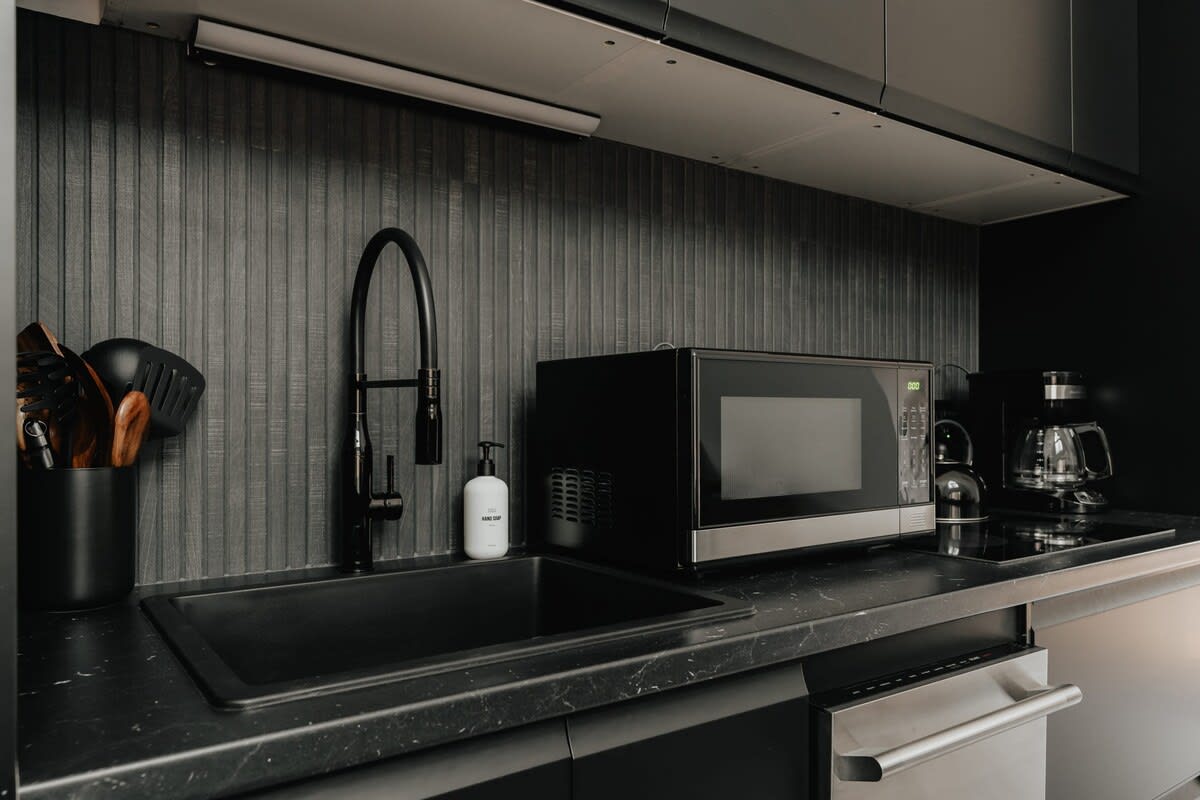 The kitchen sink area features a black undermount sink with a modern black gooseneck faucet, set against a textured black vertical slat backsplash. A microwave, kettle, and coffee maker are visible on the dark countertop.
