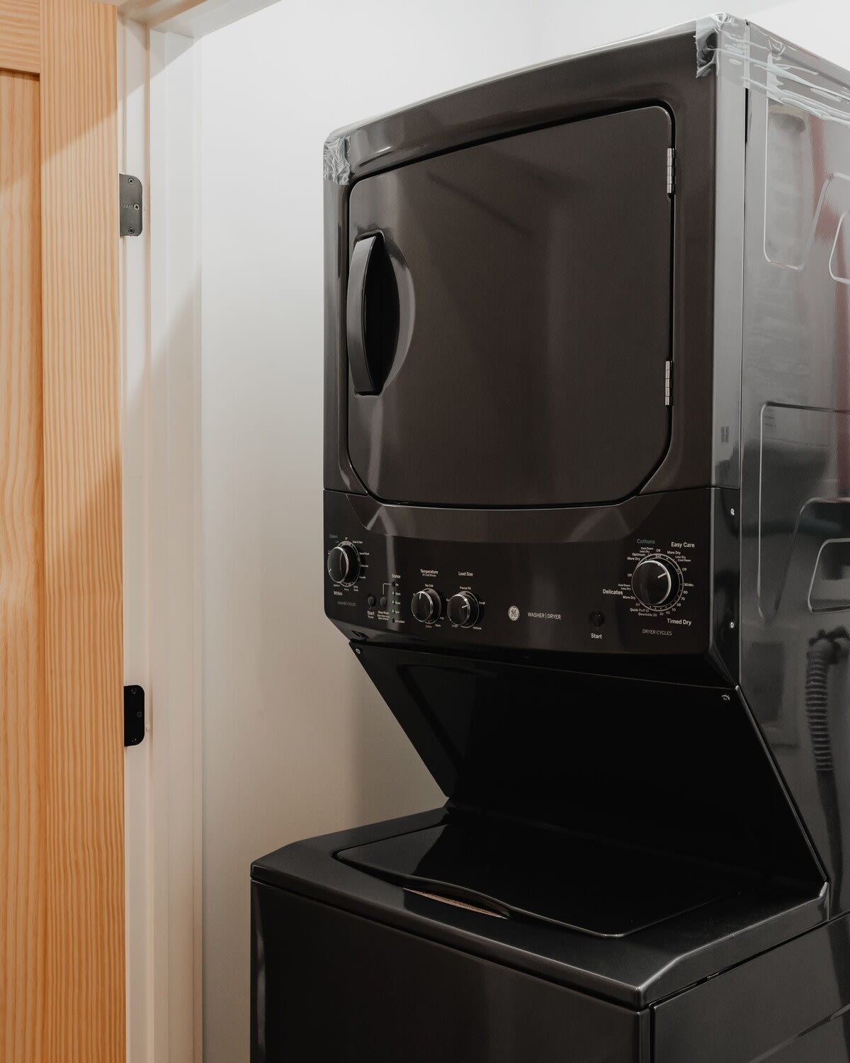 A compact laundry area featuring a black stacked washer and dryer unit with clear top lids, neatly tucked into a closet space with a light wood door.