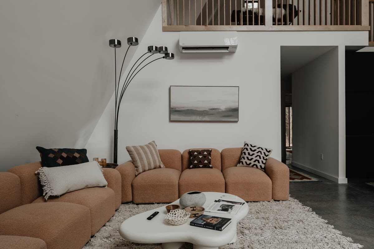 A cozy living room features a light peach sectional sofa adorned with patterned pillows, centered around a unique white organic-shaped coffee table on a plush textured rug. A multi-headed floor lamp stands beside the sofa.