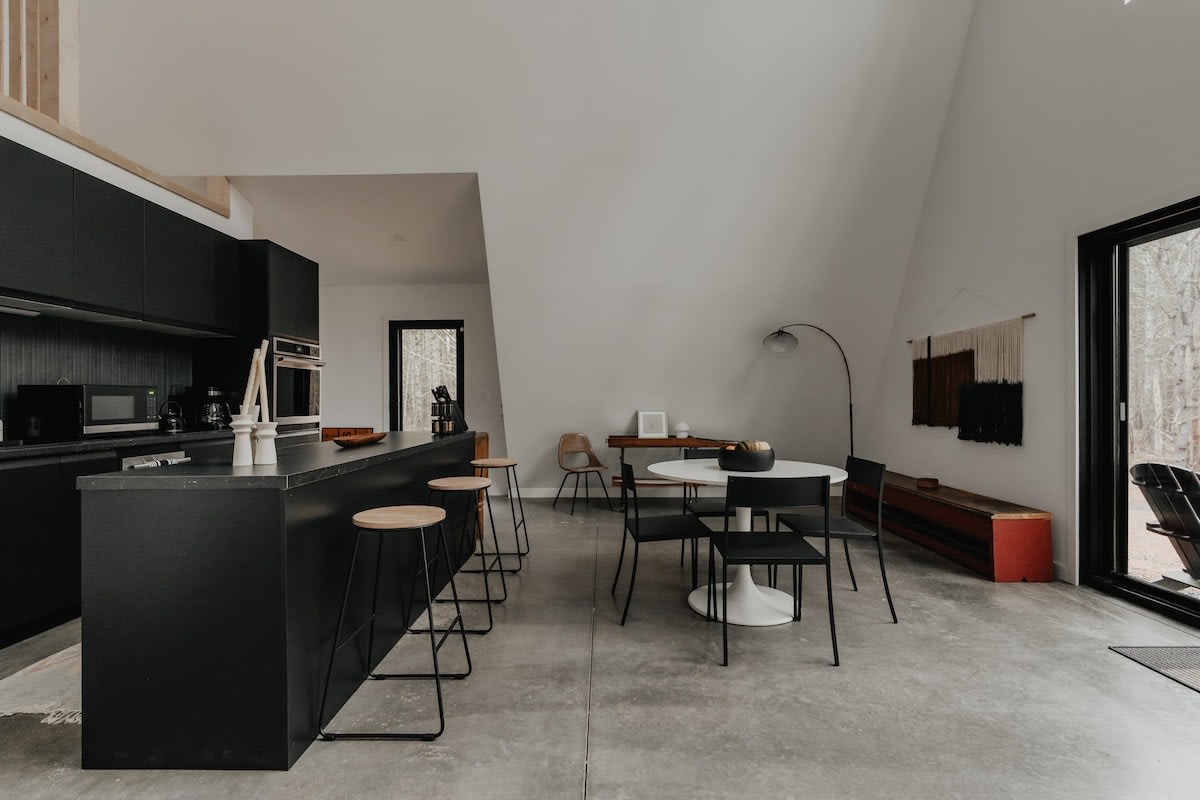 A minimalist dining area with a round white table and black chairs sits on a polished concrete floor, adjacent to a black kitchen island with bar stools. Large glass doors offer a view of the surrounding trees.