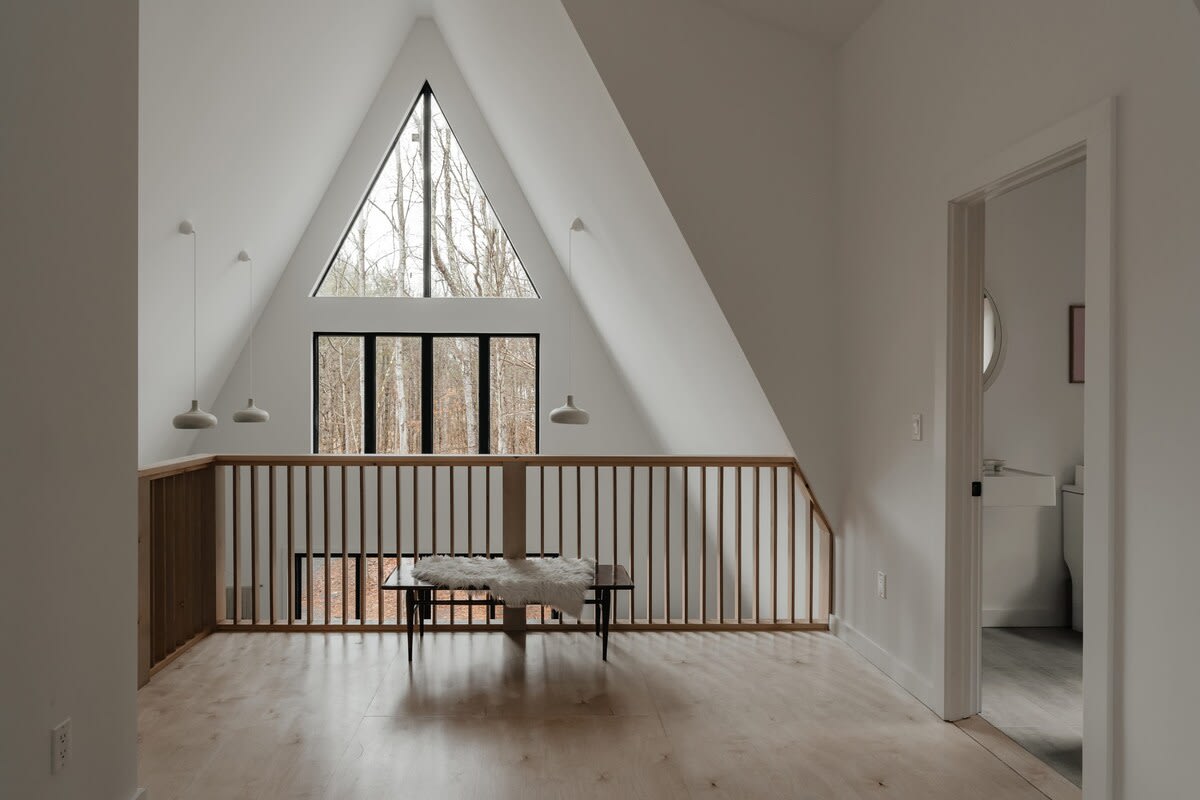 Spacious loft area with light wood flooring and a wooden railing overlooking the floor below. A large triangular window and a set of rectangular windows offer expansive views of the surrounding trees.