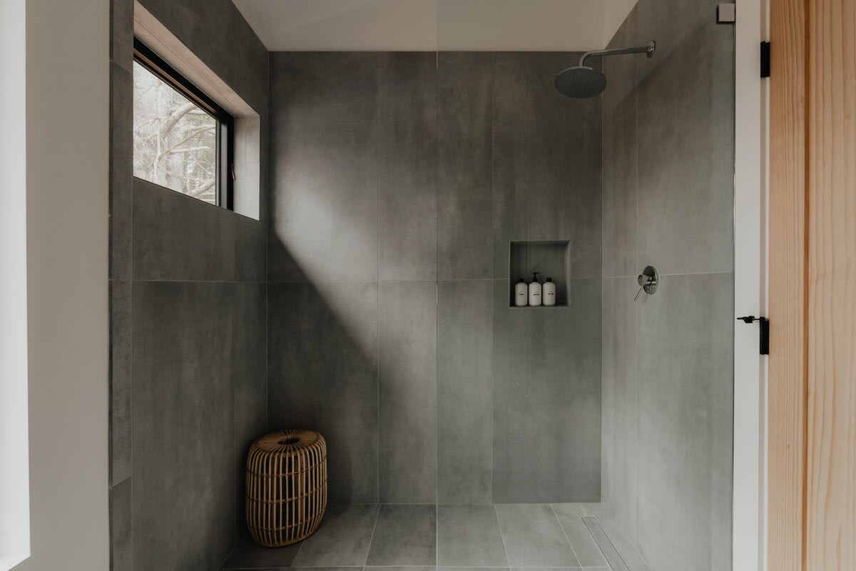 Modern walk-in shower with large grey tiled walls and floor, featuring a built-in niche for toiletries and a small wooden stool. A narrow horizontal window offers a glimpse of trees outside.