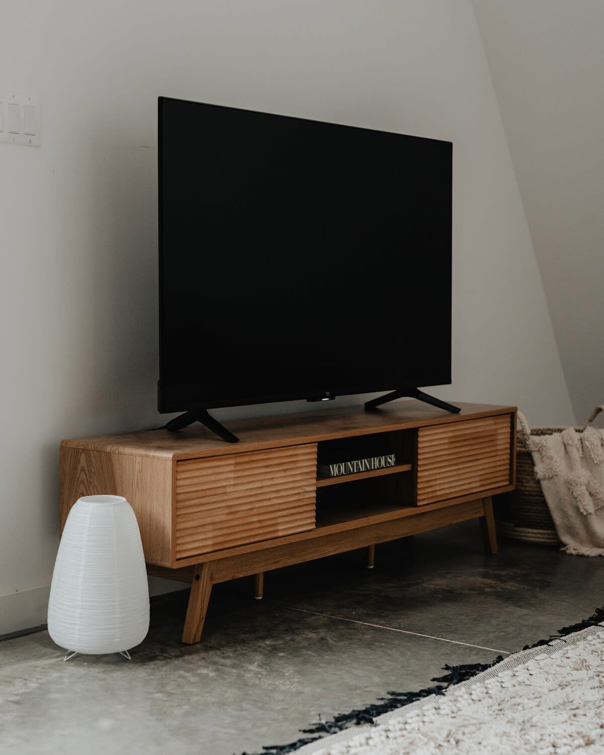 A large flat-screen television rests on a mid-century modern wooden media console with slatted doors, positioned against a white wall. A white sculptural lamp sits on the concrete floor beside it.