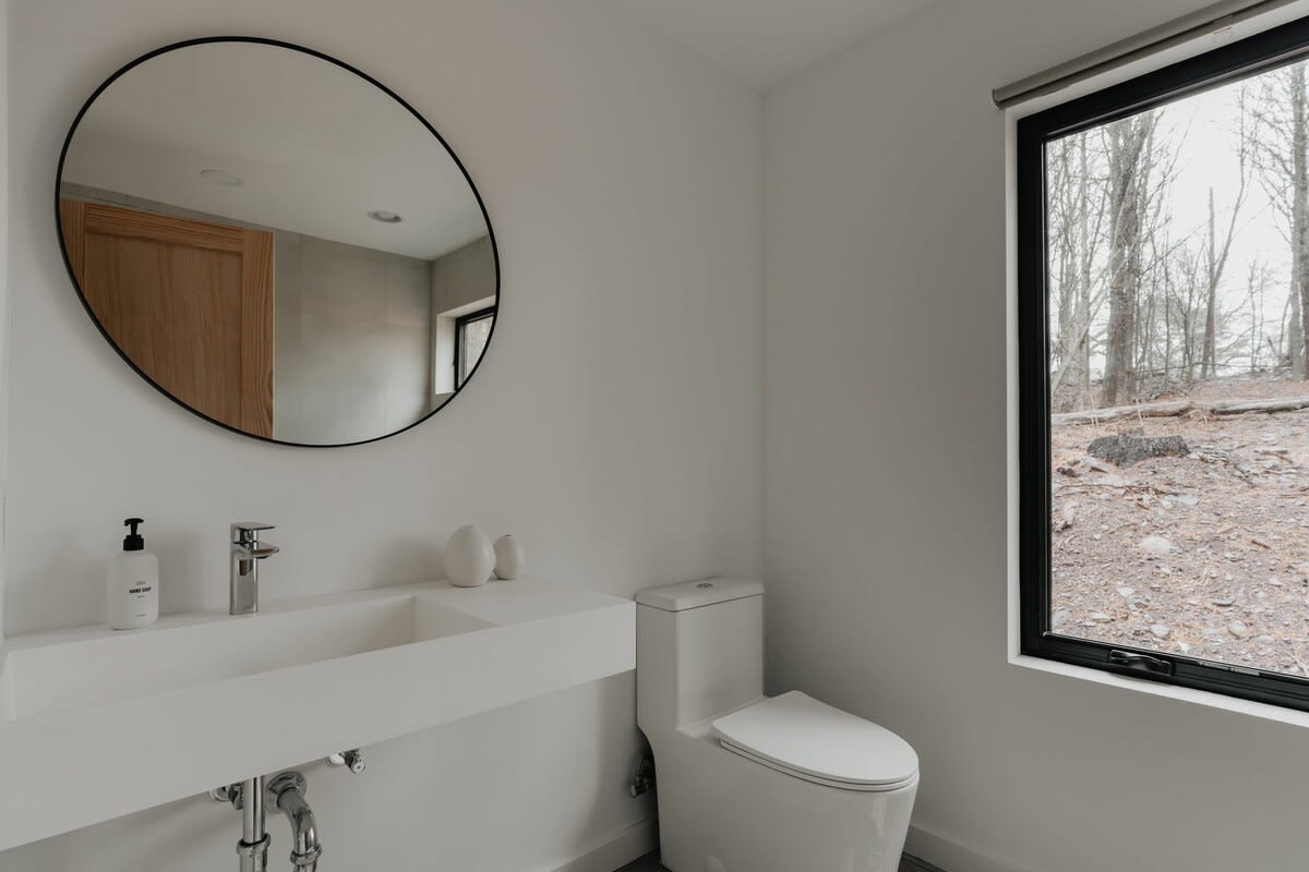 A modern bathroom featuring a white floating vanity with an integrated sink, a round black-framed mirror, a white toilet, and a large black-framed window offering a view of a sparse wooded landscape.