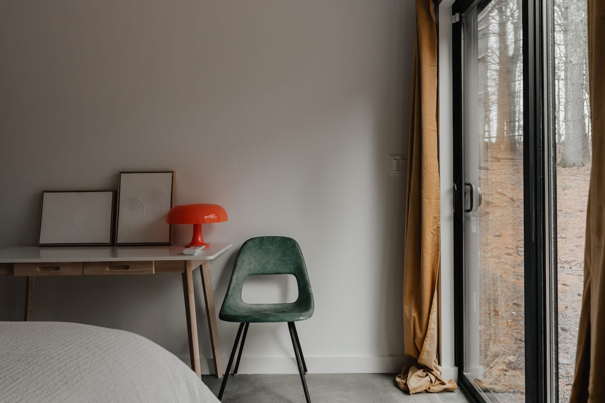 A dedicated workspace in the bedroom includes a minimalist white desk with wooden legs and a dark green modern chair. Large glass doors with a mustard yellow curtain provide natural light and a view of the forest.