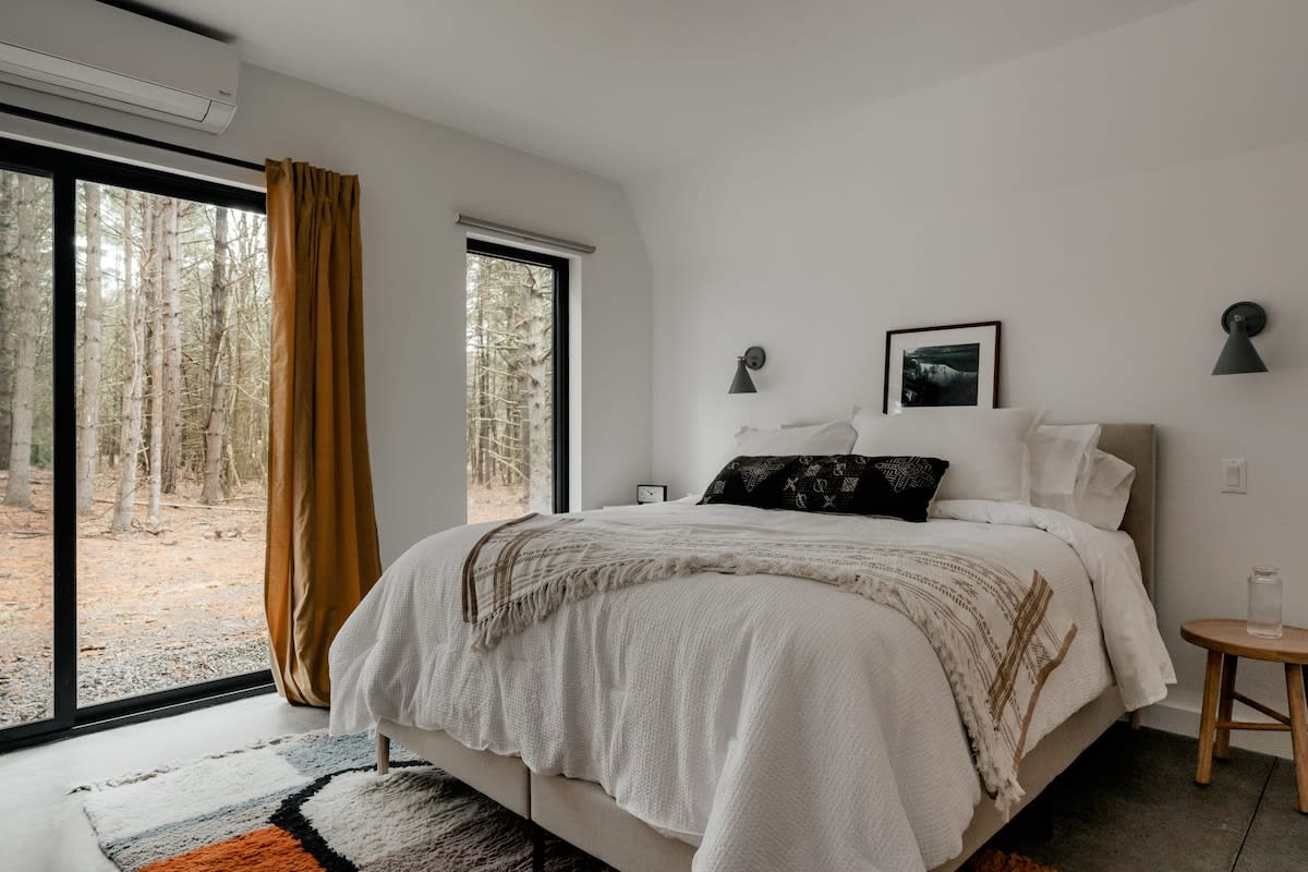 A bright bedroom features a bed with white bedding and a patterned throw, flanked by wooden side tables and wall-mounted sconces. Large sliding glass doors offer a direct view into a wooded outdoor area.