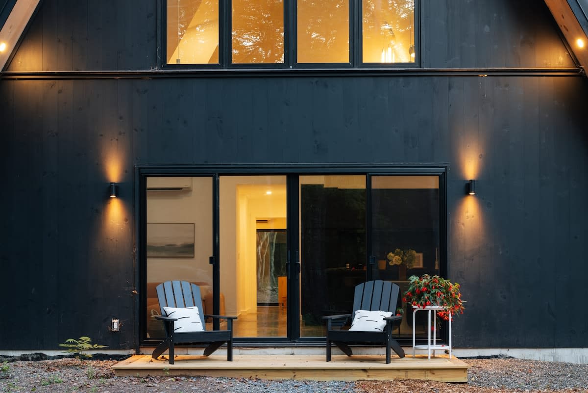 Close-up of the front deck area of the black A-frame cabin, featuring two dark Adirondack chairs with white pillows, a small side table with a potted plant, and a sliding glass door entrance, illuminated by wall sconces.