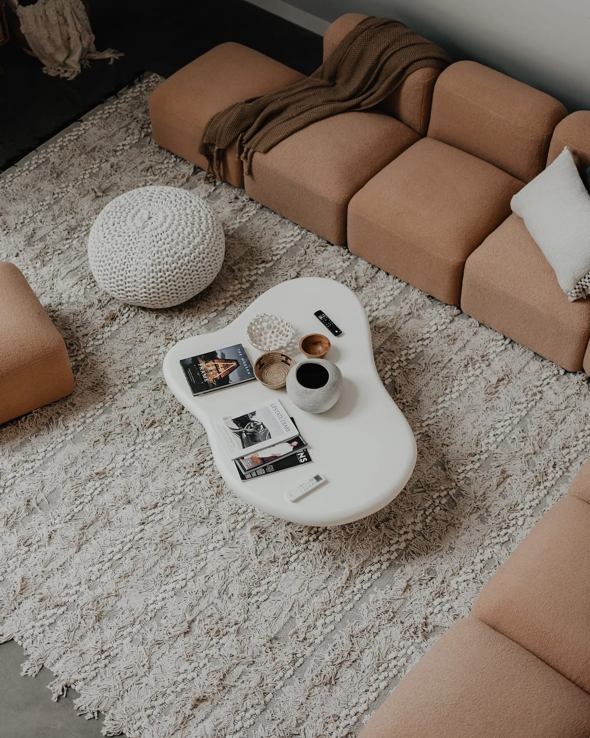 Overhead view of the living room's seating area, showcasing a light peach modular sofa, a white textured pouf, and a white organic-shaped coffee table adorned with magazines and decorative items, all on a shaggy cream rug.