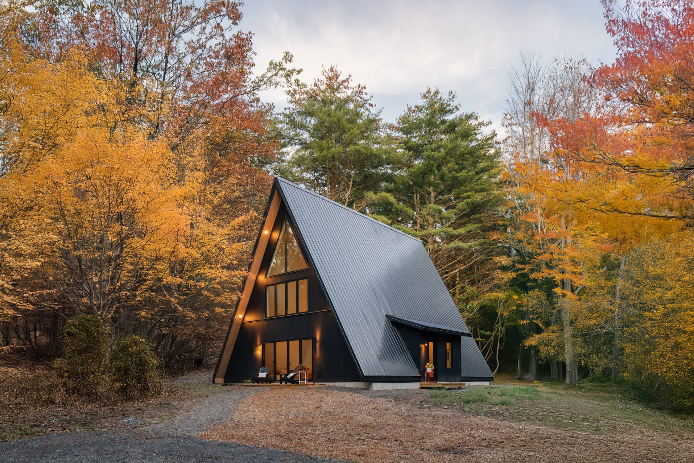 Striking black A-frame cabin surrounded by vibrant autumn foliage, with a dark metal roof and prominent triangular and rectangular windows. The property is set amidst a mix of evergreen and deciduous trees.