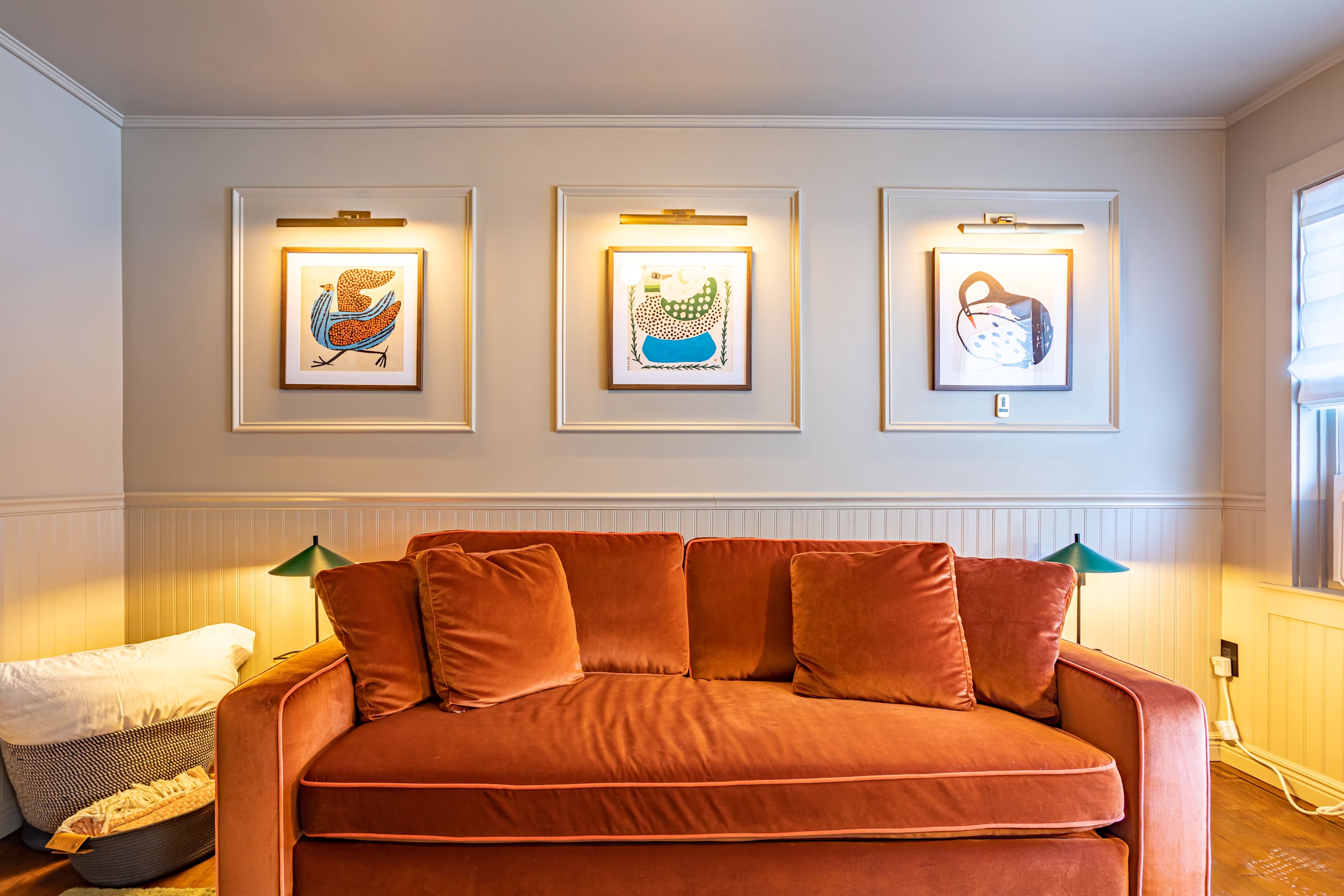 A well-lit living room featuring a rust-colored velvet sofa with multiple throw pillows, flanked by two small side tables with green lamps. Three framed art pieces with accent lighting hang above white wainscoting on the light-colored wall.