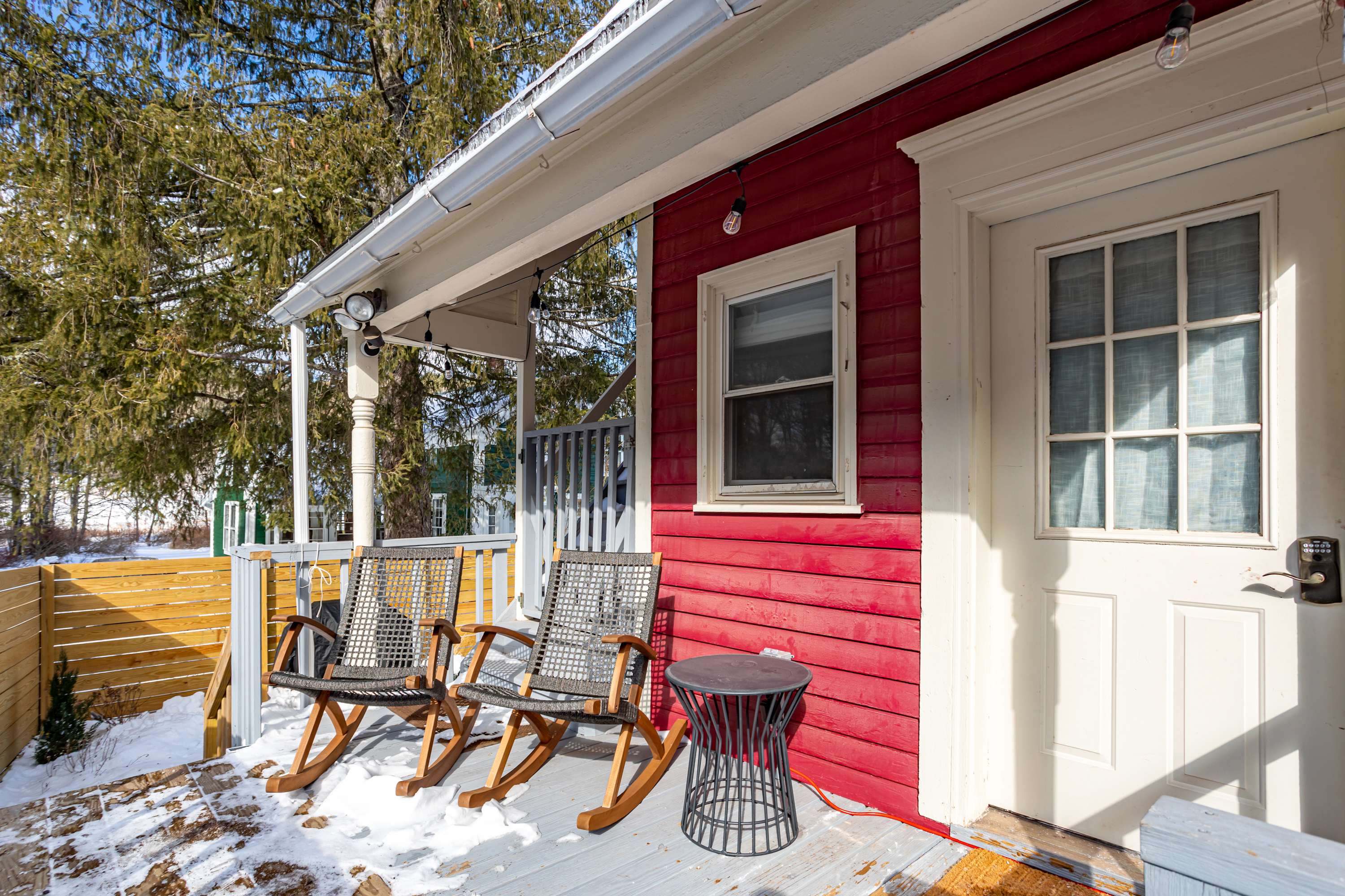 A cozy covered porch with a light gray wooden deck features two dark rocking chairs and a small black side table, set against the red siding of the house with a white door and window.