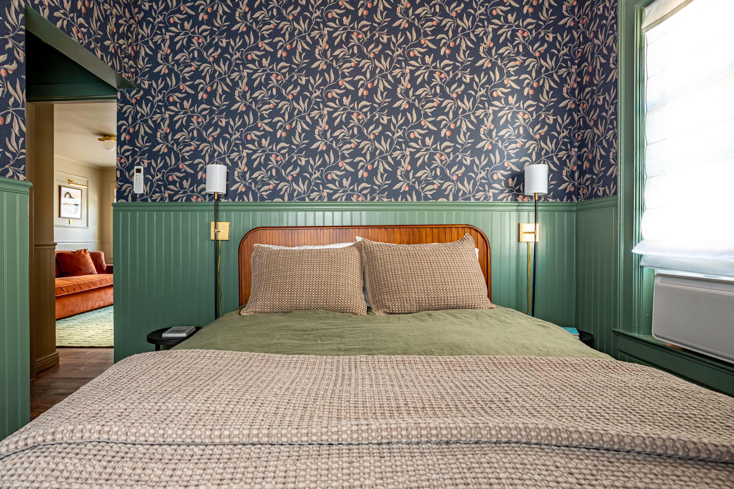 A cozy bedroom showcases dark blue botanical wallpaper above green beadboard wainscoting, featuring a bed with a light wood headboard, neutral bedding, and modern wall-mounted sconces on either side.