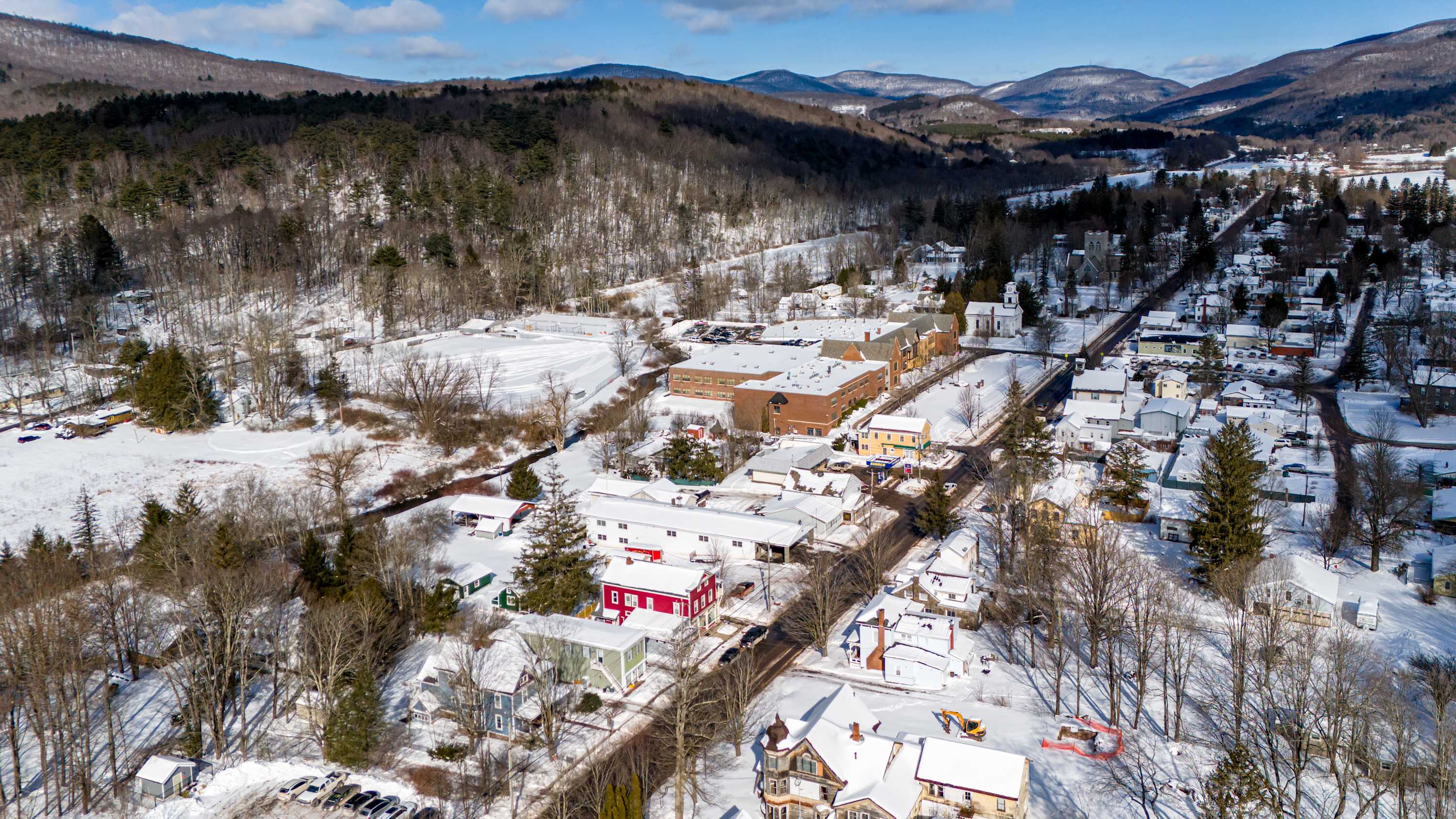 A wide aerial shot showcasing a snow-covered village nestled in a valley, surrounded by forested mountains under a bright blue sky. The red property is visible among other buildings, with a school complex in the distance.