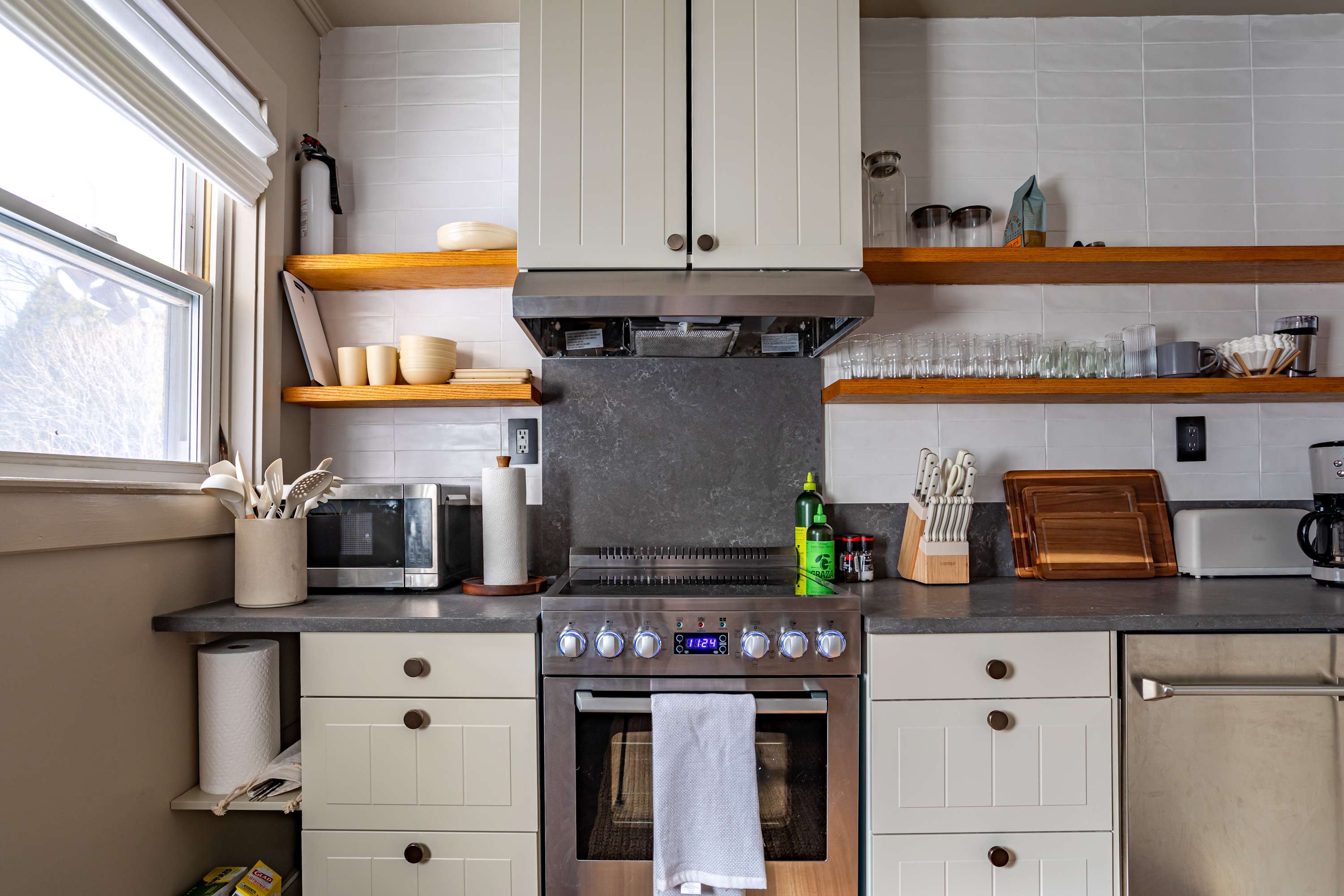 A modern kitchen with white shaker cabinets, open wooden shelving, and dark countertops features a stainless steel range with a matching hood, white subway tile backsplash, and various cooking essentials.