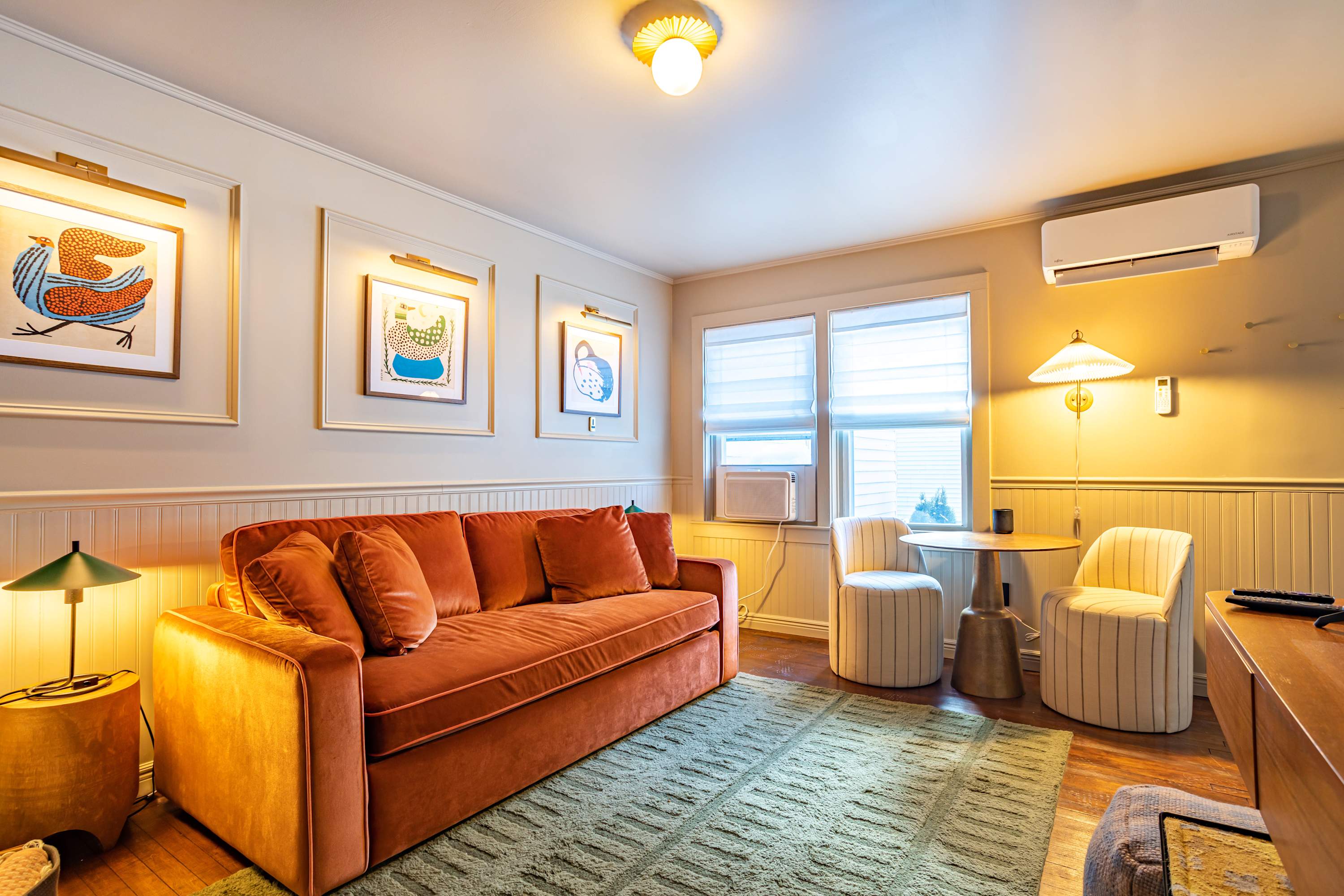 A warm living room features an orange velvet sofa, a green area rug, and white beadboard wainscoting, complemented by framed artwork with accent lighting and a small dining nook by the window.