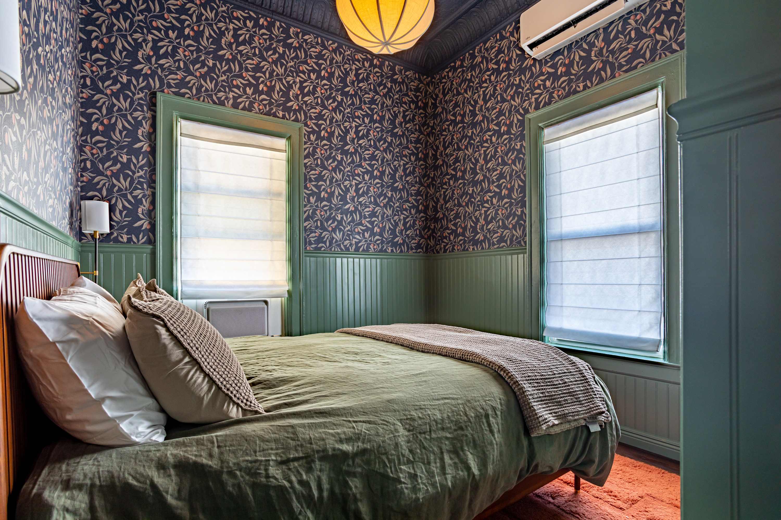 A vibrant bedroom features dark blue wallpaper with a botanical pattern and green beadboard wainscoting, housing a bed with a wooden headboard and green bedding, illuminated by a unique ceiling light fixture.