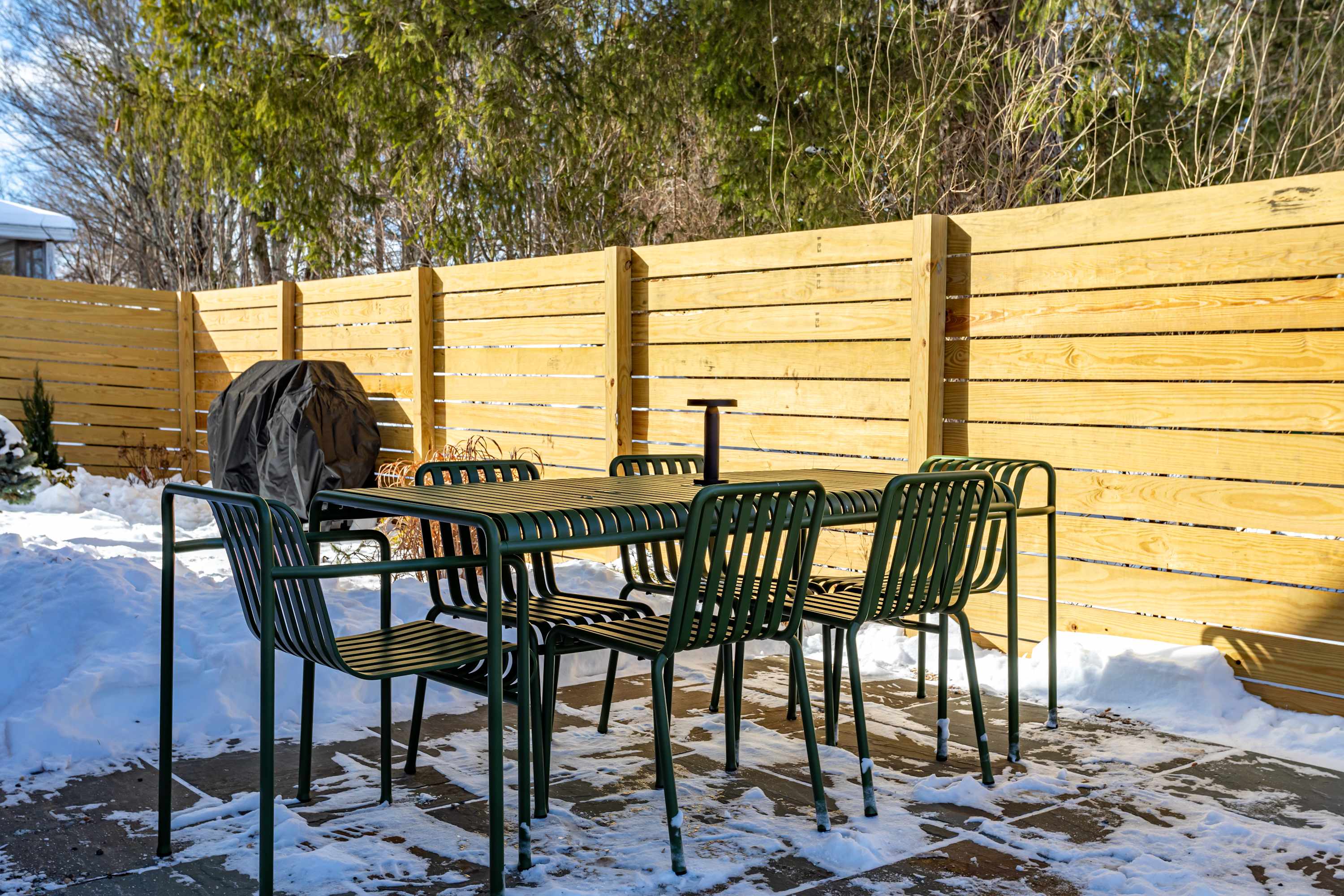 An outdoor patio area features a dark green metal dining table with six matching chairs, a covered BBQ grill, and a light wooden privacy fence, surrounded by snow-covered ground.