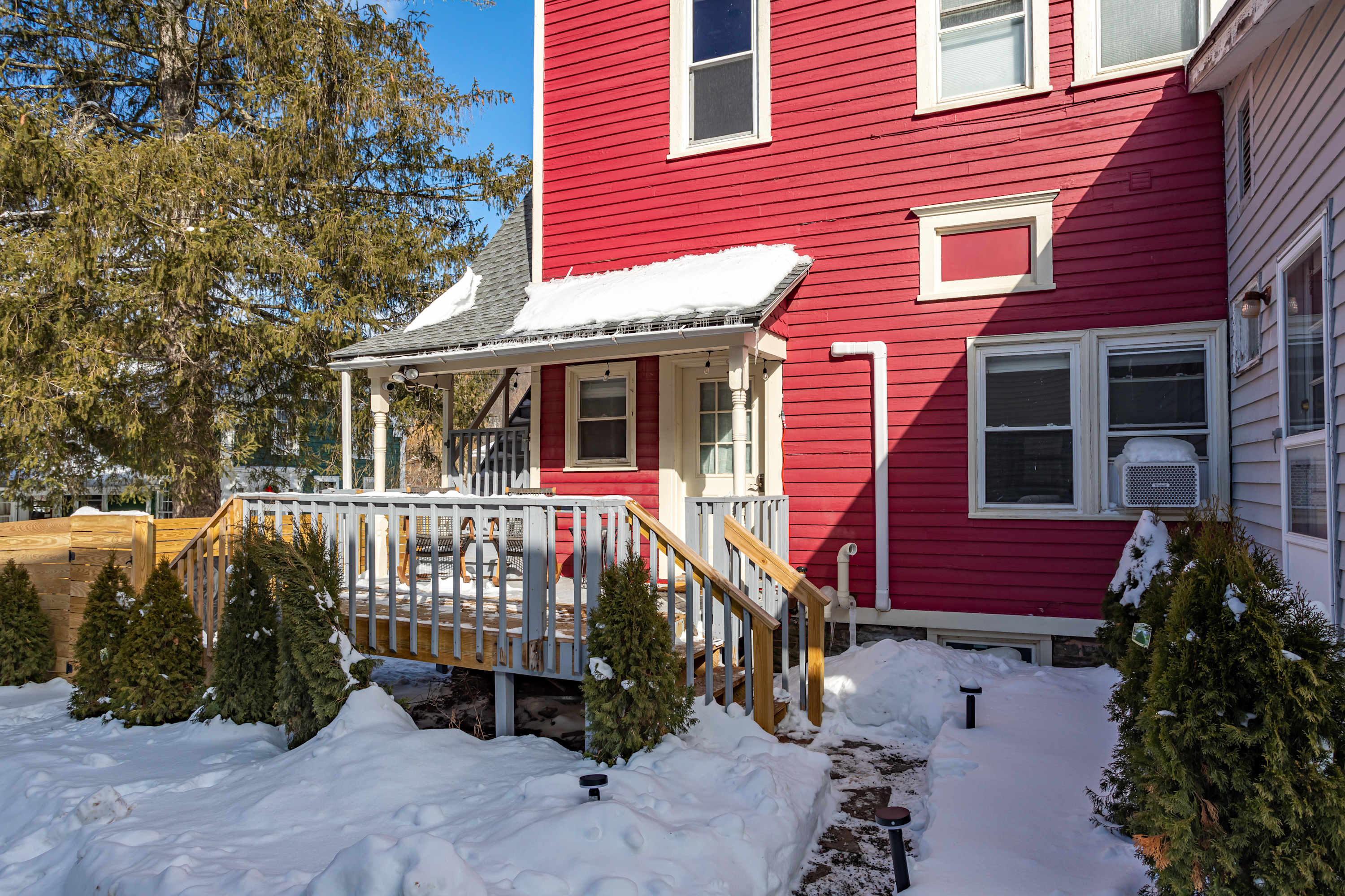 A vibrant red house with white trim features a covered porch and wooden deck with railings, surrounded by snow-covered ground and evergreen trees under a clear blue sky.