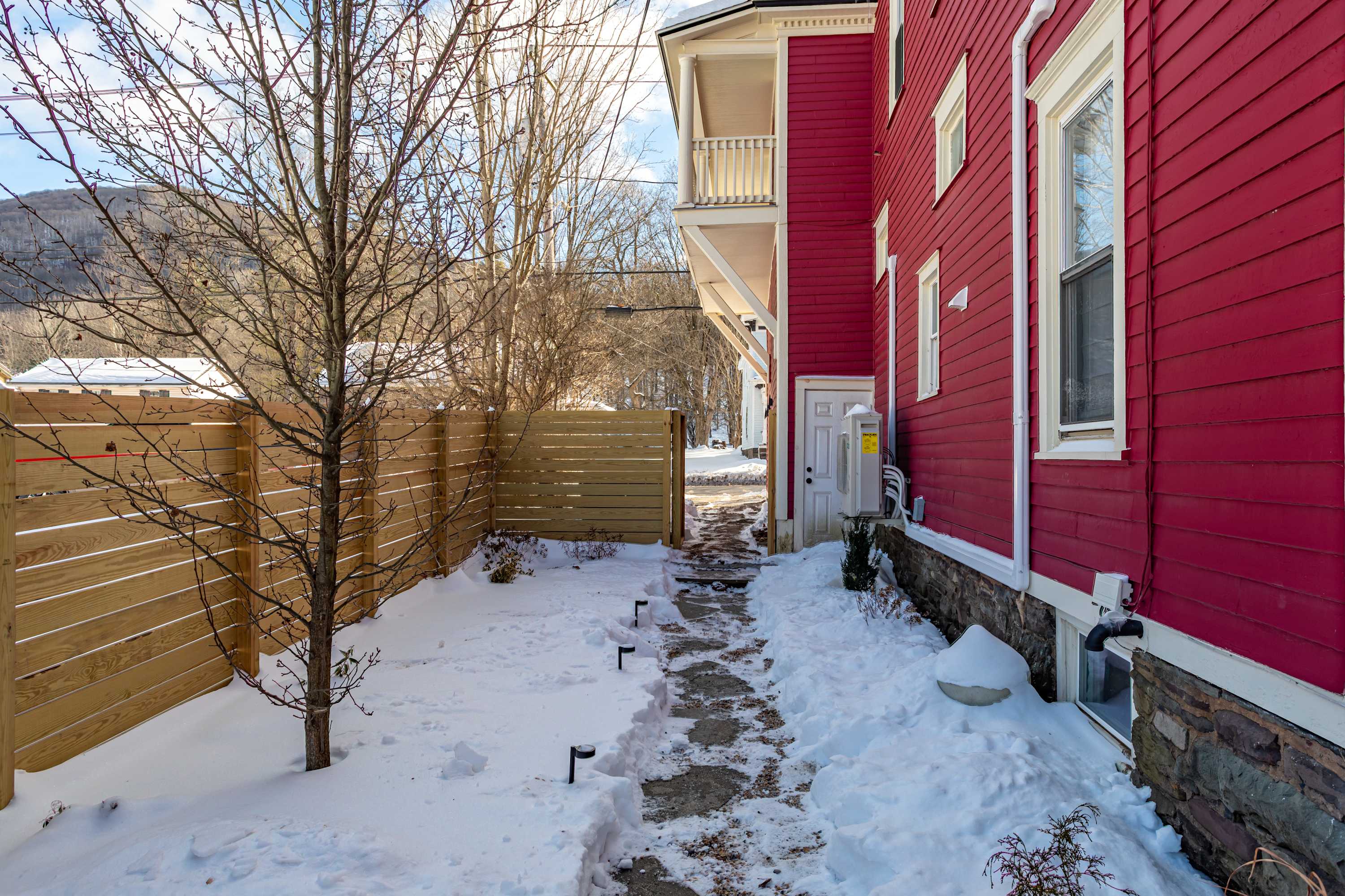 A narrow snow-covered pathway lined with small lights leads alongside the red-sided house, past a wooden privacy fence and a bare tree, towards a distant gate.