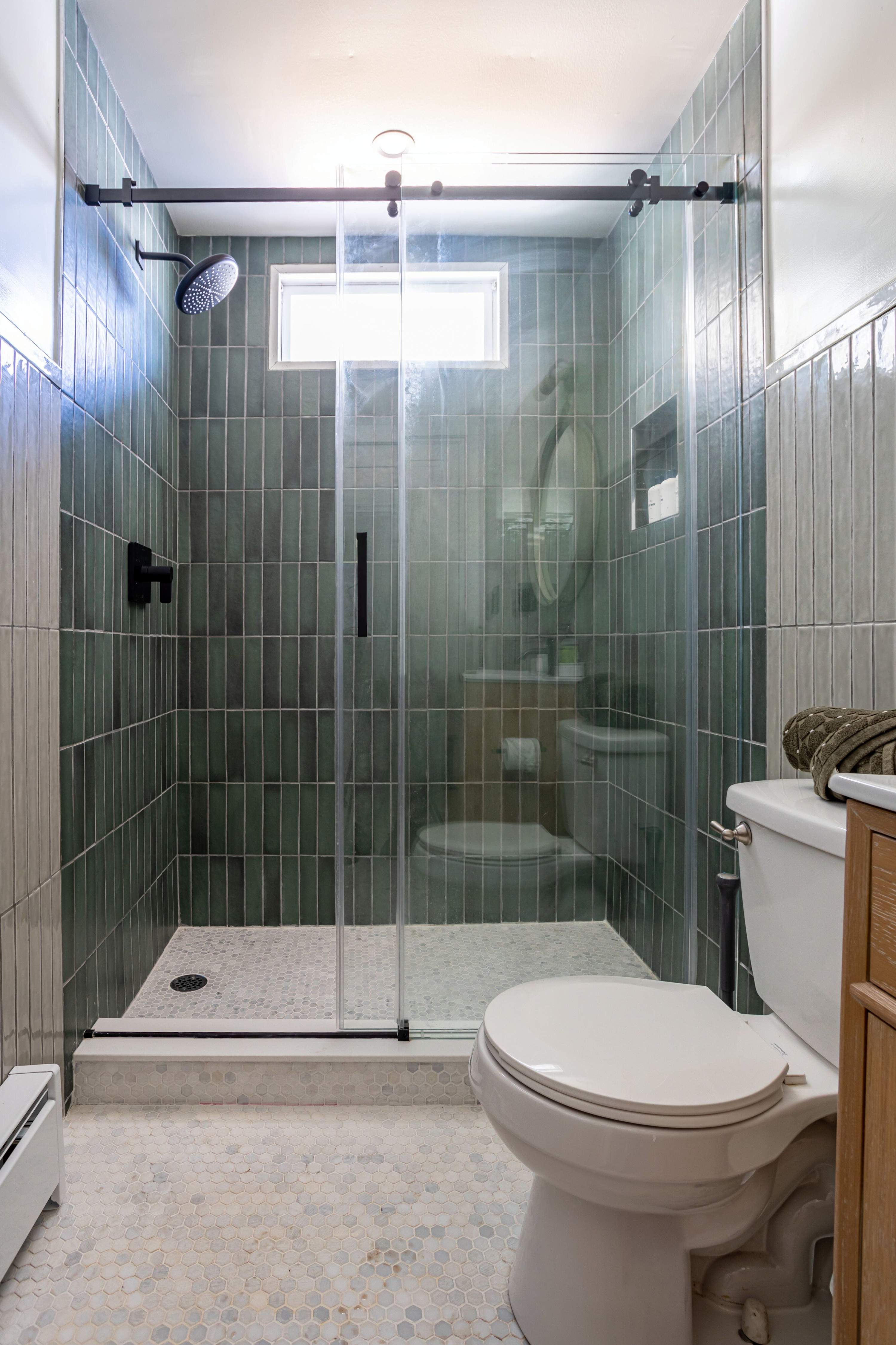 A contemporary bathroom features a walk-in shower with dark green vertical subway tiles and a glass enclosure, alongside a white toilet and light hexagonal floor tiles, creating a sleek and clean aesthetic.