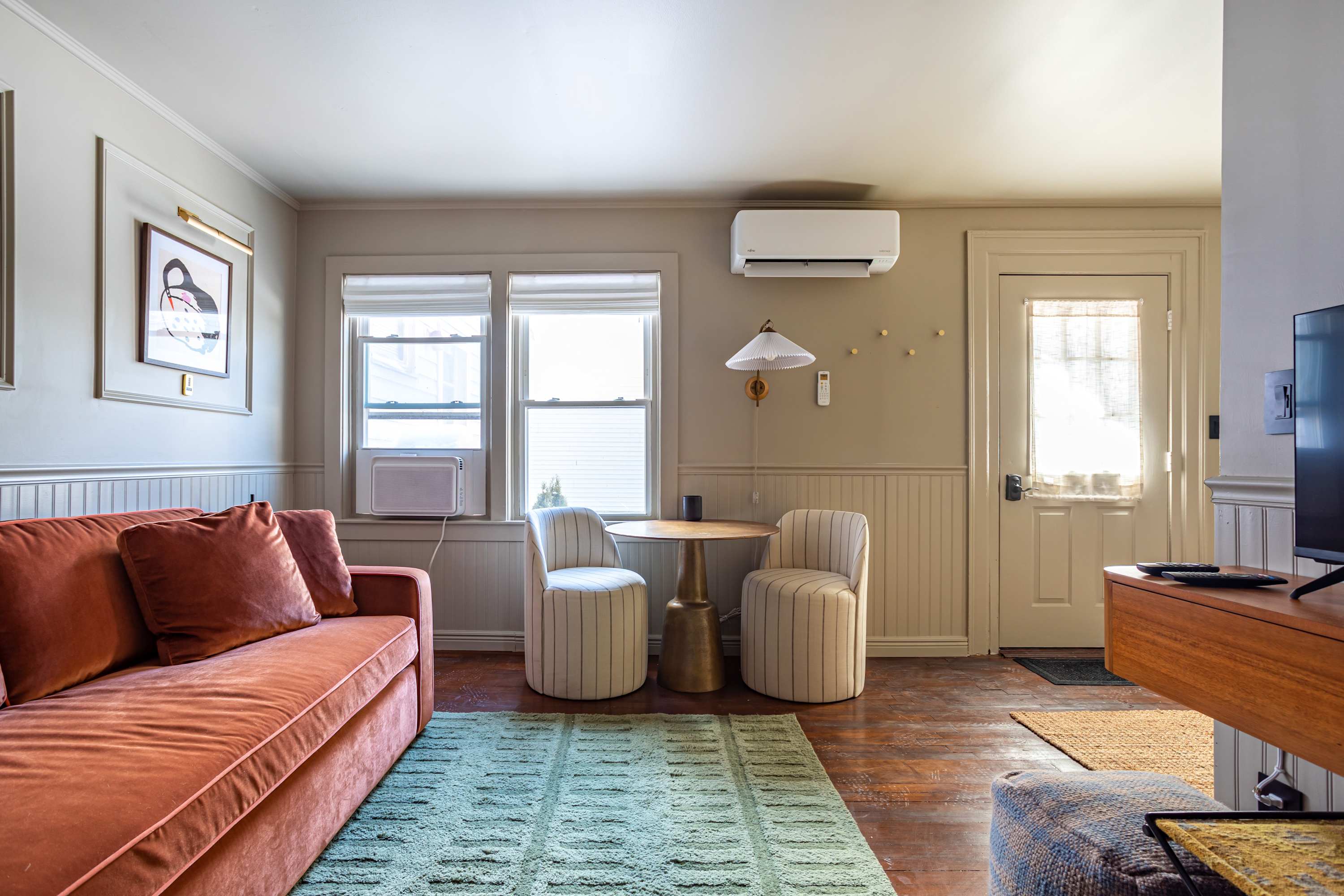 A cozy living room featuring a long coral-colored sofa, a small round dining table with two striped chairs, and hardwood floors covered by a green rug. A wall-mounted AC unit and a window AC unit are visible.