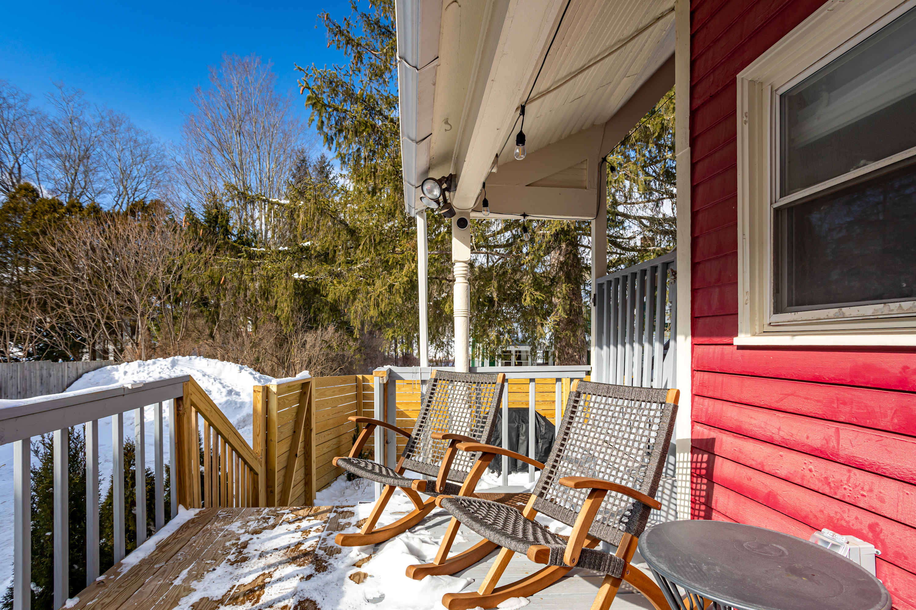 A cozy wooden porch with three modern rocking chairs and a small round table, partially covered in snow, overlooking a snowy backyard with evergreen trees under a clear blue sky. The house exterior features red siding.
