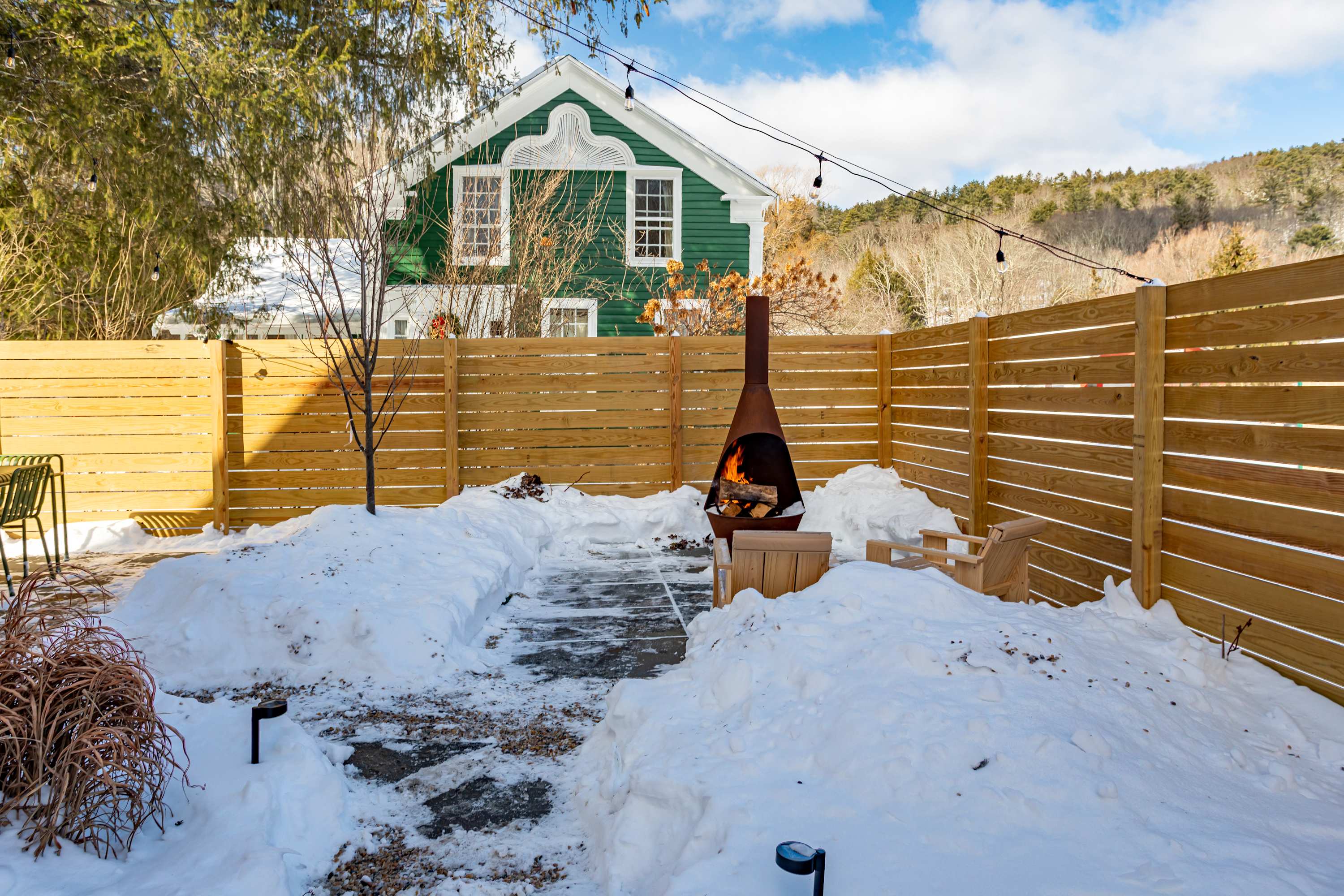 A snowy backyard enclosed by a wooden fence, featuring a tall metal fire pit with a burning fire, surrounded by snowdrifts and a stone pathway. A green house with white trim is visible in the background.