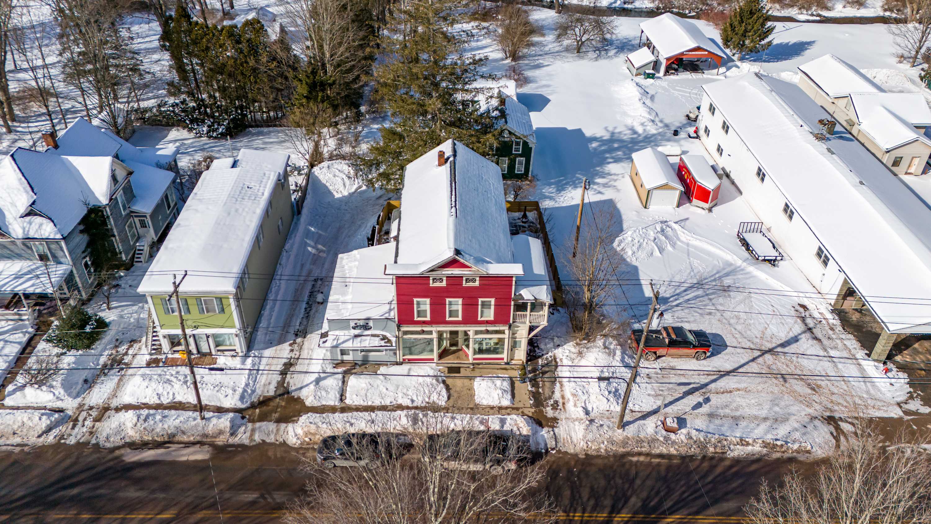 An aerial view of a red two-story house with a snow-covered roof, situated on a street with other snow-dusted homes and bare trees. A car is parked on the street in front of the house.