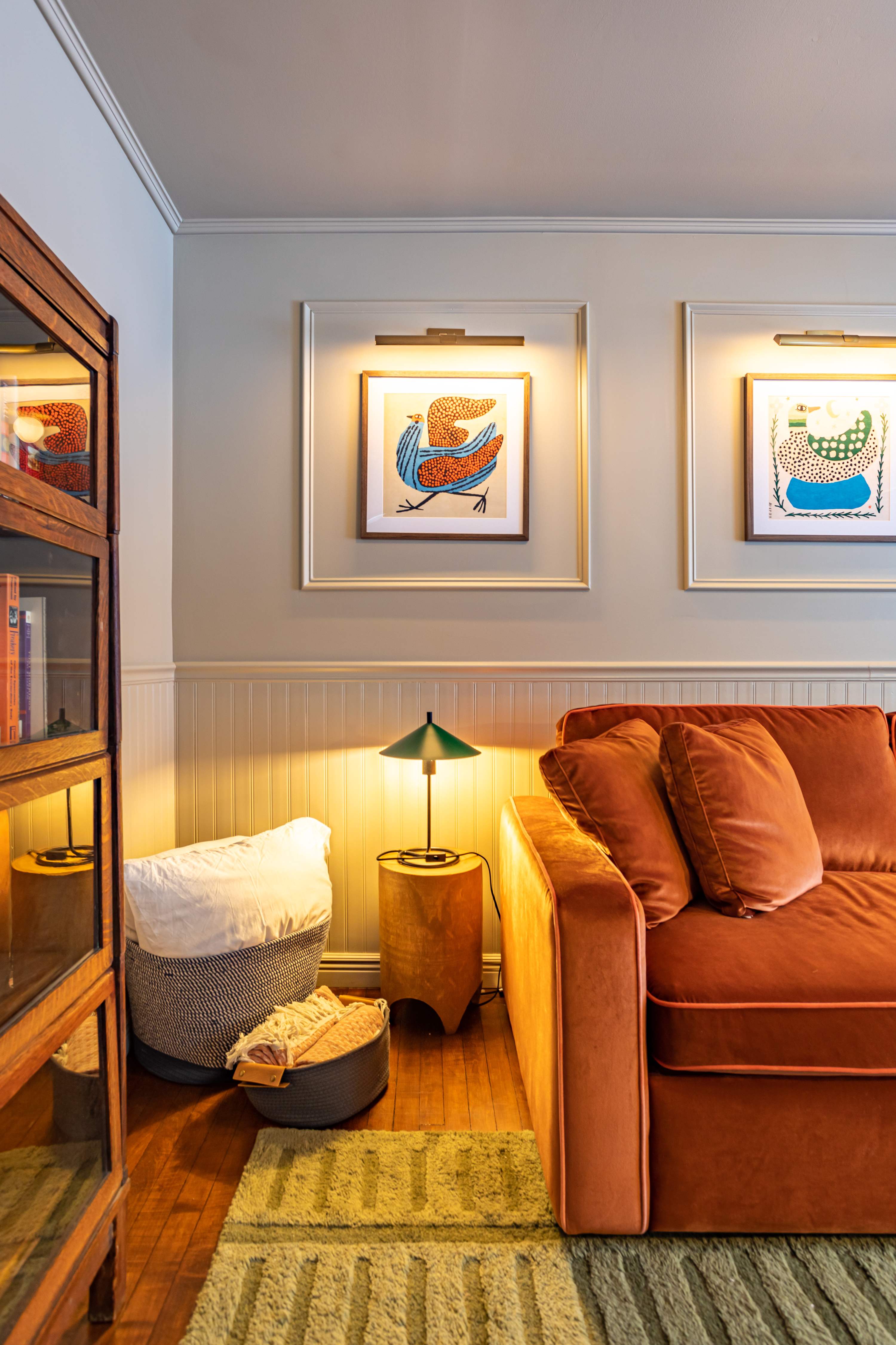 A close-up of the living room showing a rust-colored velvet sofa with throw pillows, a small wooden side table with a green lamp, and a vintage barrister bookcase. Framed artwork with accent lighting hangs above the wainscoting.