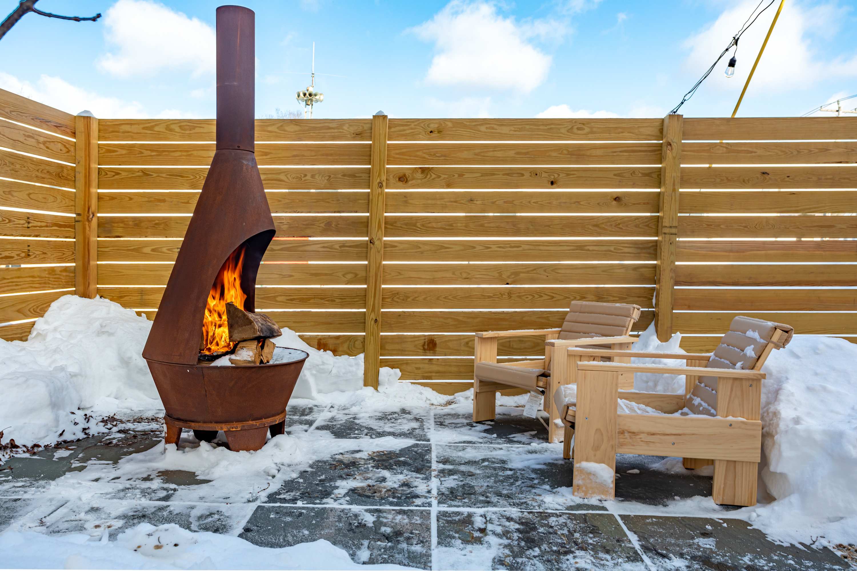 A rustic outdoor patio features a tall, conical metal fire pit with a roaring fire, flanked by two light wooden Adirondack-style chairs, all surrounded by snow and a wooden privacy fence.