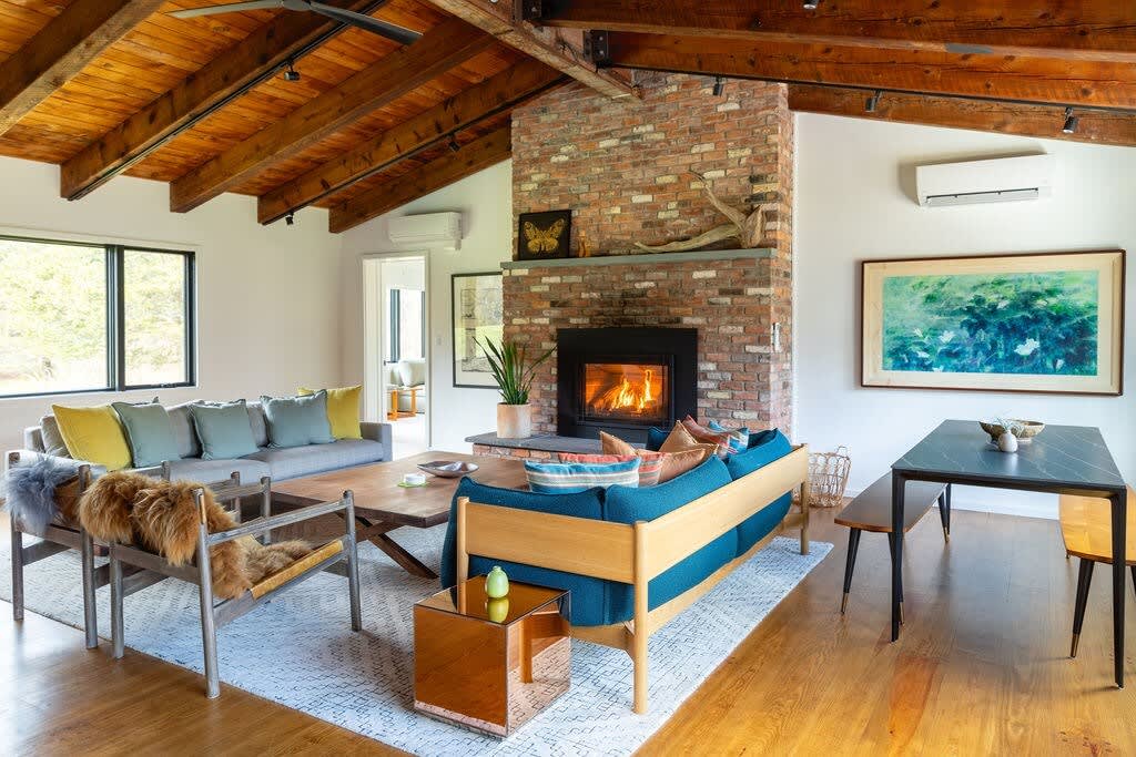 Cozy living room featuring a prominent brick fireplace, exposed wooden beam ceiling, light blue and grey sofas, and a small dining table with benches, all on hardwood floors.