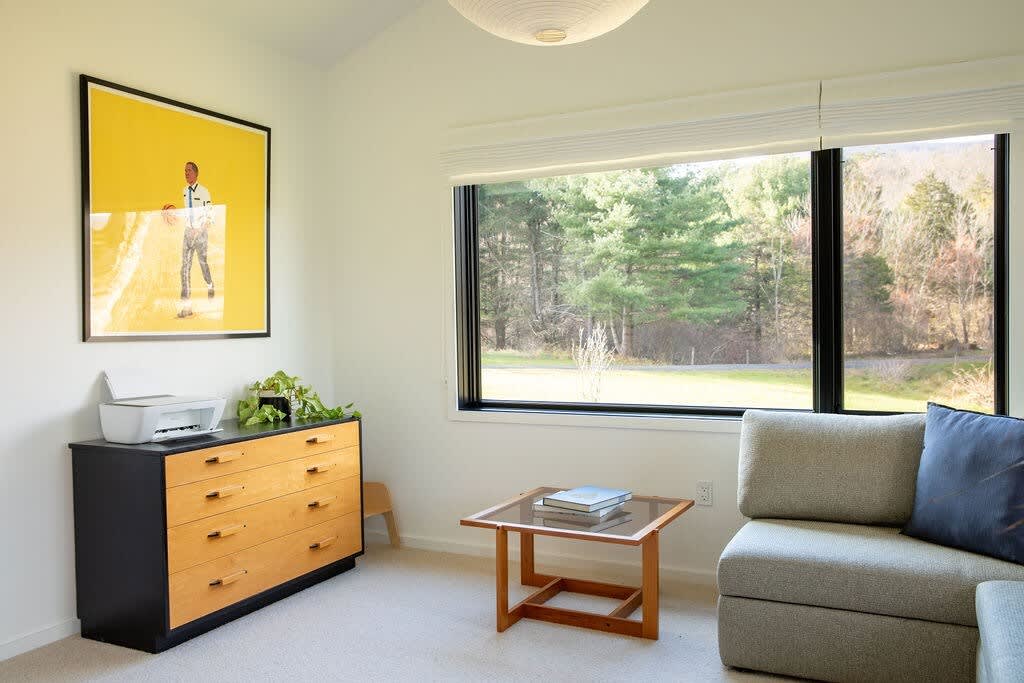Corner of a bright bedroom featuring a large window with green outdoor views, a wooden dresser with a printer, a comfortable sofa, and a small coffee table, suitable for work or relaxation.
