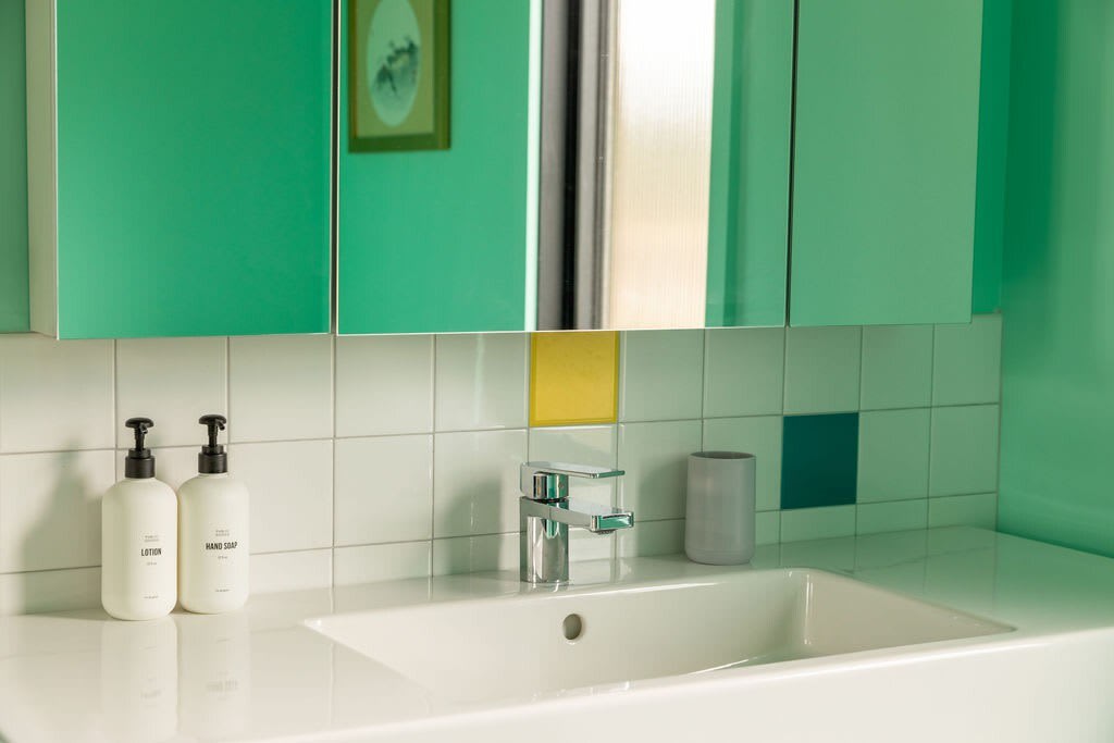 A close-up of the bathroom sink area, featuring a white vanity with a chrome faucet, white square wall tiles with colorful accent squares (yellow, blue), and a bright teal wall above.