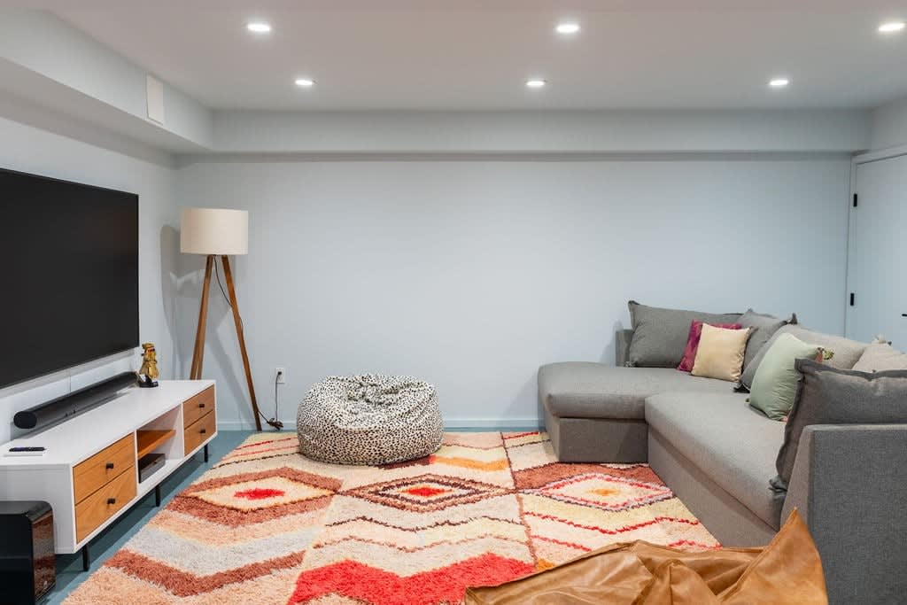 A cozy media room features a large wall-mounted TV above a white and wood media console, with a grey sectional sofa and a patterned area rug. Recessed lighting brightens the light blue walls, creating a comfortable space for entertainment.