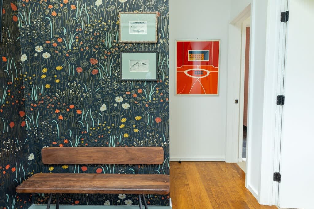 A vibrant hallway or foyer features dark floral wallpaper with intricate patterns, a rustic wooden bench, and framed artwork. The space is illuminated by natural light, with warm hardwood floors leading to a white doorway.