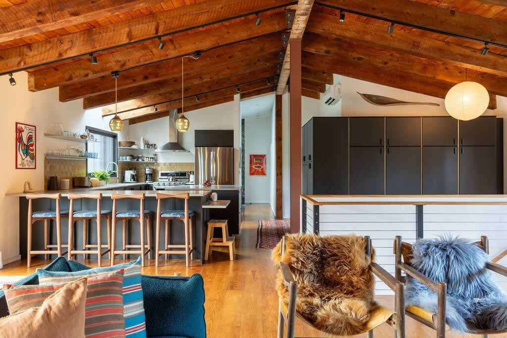 Open-concept kitchen and living area with exposed wooden beams, a kitchen island with bar stools, stainless steel appliances, and a view into a hallway, all on hardwood floors.