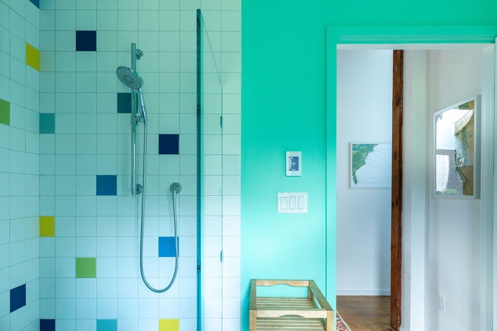 A shower area with white square tiles accented with colorful blue, yellow, and green squares, and a bright teal wall. A showerhead and a wooden bench are visible inside the shower.