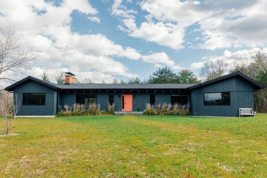 The front exterior of a dark gray modern house with a vibrant orange front door, set against a bright blue sky with white clouds. The house is surrounded by a green lawn and ornamental grasses, with trees in the background.